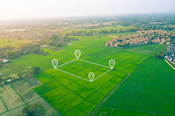 Aerial view of surveyed farmland on the edge of a town, with pin markers and dotted lines plotting out a block, like a parcel of land waiting to be developed.
