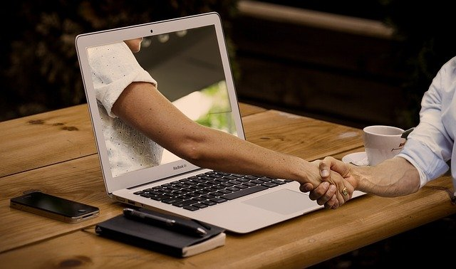 A hand reaches out of a laptop screen on a wooden desk to shake hands with someone in person, representing a digital connection becoming a real one.